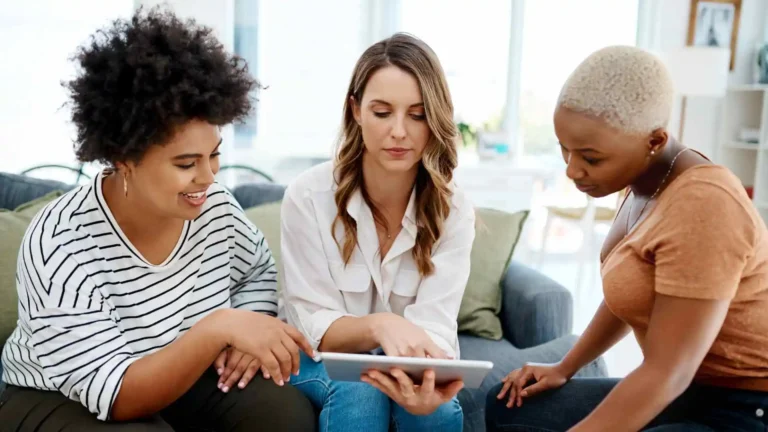 Três mulheres conversam e analisam dados em um tablet durante uma avaliação psicossocial.